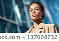 Confident young woman wearing glasses and beige blazer, standing near modern glass architecture in natural daylight. 137008732