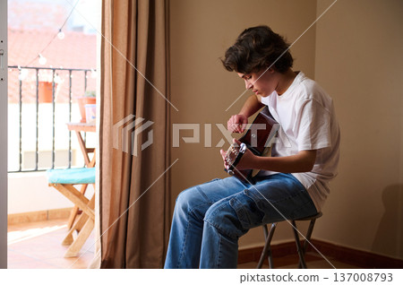 Teen Boy Playing Acoustic Guitar Indoors on Stool by Sunlit Balcony Teen Boy Playing Acoustic Guitar Indoors on Stool by Sunlit Balcony 137008793