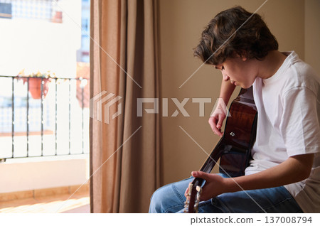 Teen Boy Playing Acoustic Guitar Indoors by Sunlit Balcony in Casual White T-Shirt Teen Boy Playing Acoustic Guitar Indoors by Sunlit Balcony in Casual White T-Shirt 137008794