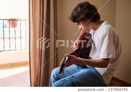 Teenage Boy Playing Acoustic Guitar Indoors by Sunlit Balcony Window 137008795