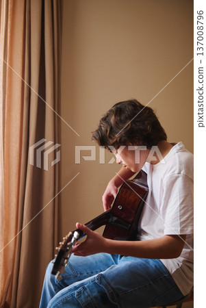 Teen Boy Playing Acoustic Guitar at Home by a Window, Practicing Music and Concentration 137008796