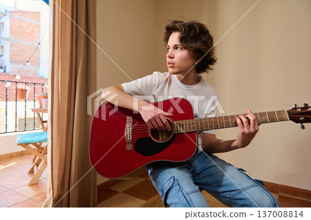 Teen Boy Playing Red Acoustic Guitar Indoors Near Balcony Window During Casual Practice 137008814