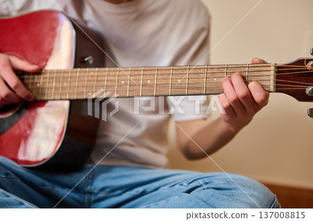 Young Person Playing Red Acoustic Guitar Sitting Cross-Legged Indoors Practicing Chords Young Person Playing Red Acoustic Guitar Sitting Cross-Legged Indoors Practicing Chords 137008815