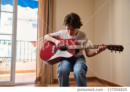 Teen Boy Playing Red Acoustic Guitar Seated Indoors Near Balcony Door Practicing Music Teen Boy Playing Red Acoustic Guitar Seated Indoors Near Balcony Door Practicing Music 137008820