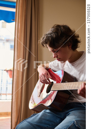 Teen Boy Playing Acoustic Guitar Indoors Near Window Practicing Music at Home 137008828