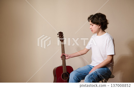 Teen Boy Sitting With Acoustic Guitar Practicing Music in Casual White T-Shirt and Jeans Teen Boy Sitting With Acoustic Guitar Practicing Music in Casual White T-Shirt and Jeans 137008852