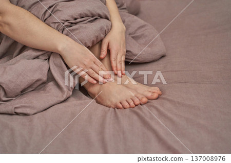 Female adult hands touching feet on bed with soft beige sheets and linen blanket for relaxation 137008976