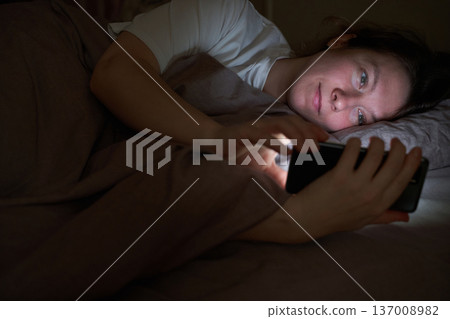 Caucasian female adult lying in bed using smartphone at night for relaxation or connection Caucasian female adult lying in bed using smartphone at night for relaxation or connection 137008982