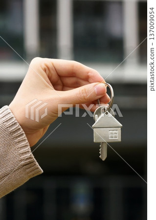 Vertical Closeup Woman Holds House Key, Apartment Building Stands In Background. Homeownership, Real Estate Deal, Purchase, Moving, Investment. Sale Buy Apartment, Mortgage, First Homeowner, New Home 137009054