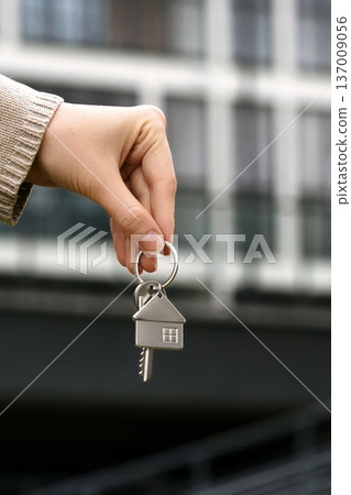Vertical closeup Woman Holds House Shaped Key, Real Estate Agent Facilitates Transfer. Modern Apartment Building, Ownership, Purchase, Investment, Relocation. Sale Buy Apartment. 137009056
