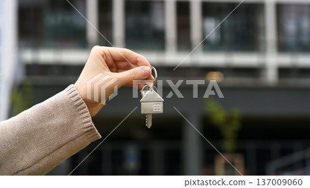 Woman Holds House Key, Apartment Building Stands In Background. Homeownership, Real Estate Deal, Purchase, Moving, Investment. Sale Buy Apartment, Mortgage, First Homeowner, New Home. Copy Space. Woman Holds House Key, Apartment Building Stands In Background. Homeownership, Real Estate Deal, Purchase, Moving, Investment. Sale Buy Apartment, Mortgage, First Homeowner, New Home. Copy Space. 137009060