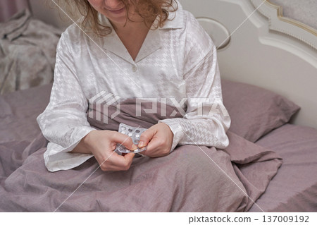 Caucasian female adult in pajamas sitting in bed holding medication blister pack in a cozy room 137009192
