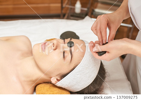 Young Caucasian Female Enjoying Relaxing Spa Treatment With Hot Stone Therapy For Face Rejuvenation Young Caucasian Female Enjoying Relaxing Spa Treatment With Hot Stone Therapy For Face Rejuvenation 137009275