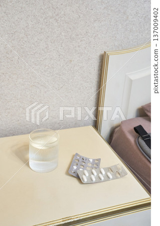 Glass of water and empty medicine blisters on nightstand beside bed with beige headboard in bedroom Glass of water and empty medicine blisters on nightstand beside bed with beige headboard in bedroom 137009402