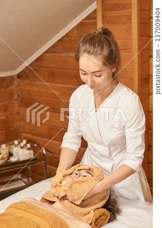 Female Adult Providing Relaxing Spa Treatment To Young Caucasian Woman In Serene Wooden Environment 137009404