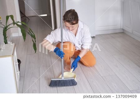 Caucasian female adult sweeping floor in kitchen wearing gloves for cleaning and household chores Caucasian female adult sweeping floor in kitchen wearing gloves for cleaning and household chores 137009409