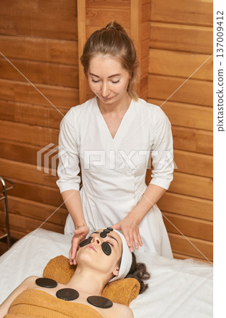Female Spa Therapist Performing Hot Stone Therapy On Young Caucasian Woman In Relaxing Wellness Female Spa Therapist Performing Hot Stone Therapy On Young Caucasian Woman In Relaxing Wellness 137009412
