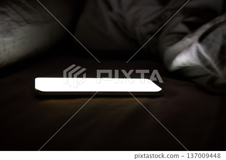 Glowing smartphone on bed with dim lighting, illuminating darkness and creating eerie ambience Glowing smartphone on bed with dim lighting, illuminating darkness and creating eerie ambience 137009448
