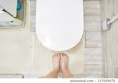 Female feet on bath mat facing toilet in modern bathroom with minimalist decor and light tiles Female feet on bath mat facing toilet in modern bathroom with minimalist decor and light tiles 137009478