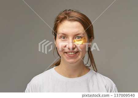 Caucasian young adult female smiling with gold under-eye patch for skincare and wellness routine Caucasian young adult female smiling with gold under-eye patch for skincare and wellness routine 137009540