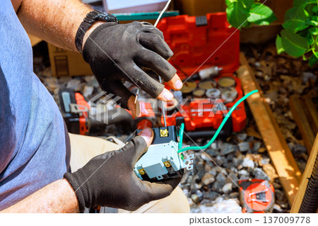 Worker connects wires to device while using tools in workshop area filled with supplies 137009778