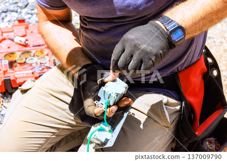 Electrician repairs electrical component using tools while seated on ground outdoors 137009790