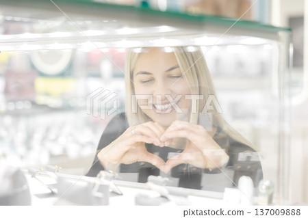 Woman making heart shape with hands while admiring jewelry in shop window Woman making heart shape with hands while admiring jewelry in shop window 137009888