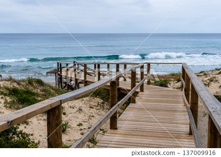 Wooden boardwalk leading to the beach in Menorca on a cloudy day. Wooden boardwalk leading to the beach in Menorca on a cloudy day. 137009916