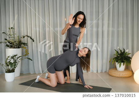 Yoga practice in a studio with two women demonstrating a pose during a morning class on a soft mat with plants in the background 137010015