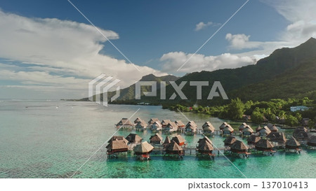 Tropical island landscape featuring luxury overwater bungalows extending into the clear turquoise lagoon, surrounded by lush green mountains and a beautiful blue sky Tropical island landscape featuring luxury overwater bungalows extending into the clear turquoise lagoon, surrounded by lush green mountains and a beautiful blue sky 137010413