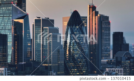 United Kingdom, London: Illuminated London skyline at sunset featuring iconic skyscrapers like the Gherkin and modern architecture of the financial district. Drone flight 137010415