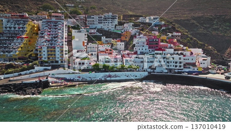 Tenerife: coastal modern colorful buildings hotels resort town on steep hillside, overlooking the Atlantic Ocean with sparkling water under a bright sun, inviting vacation and tourism. Aerial panorama 137010419