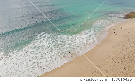 Aerial drone view shows surfers and people enjoying the turquoise water and sandy beach, with gentle waves breaking on the shore, experiencing tropical vacation and summer fun in Bali Aerial drone view shows surfers and people enjoying the turquoise water and sandy beach, with gentle waves breaking on the shore, experiencing tropical vacation and summer fun in Bali 137010430