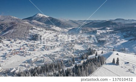 Aerial view of charming mountain village, featuring snow-covered houses nestled among pine trees, creating serene winter landscape, clear blue sky. Bukovel ski resort. Winter nature travel background 137010432