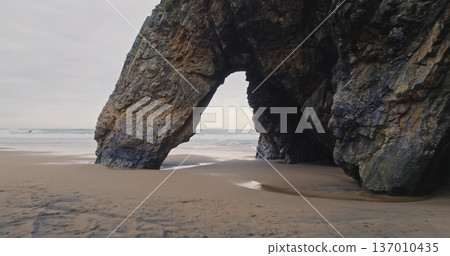 Portugal, Lisbon: Natural rock arch on Praia da Adraga sand beach, creating dramatic coastal landscape with ocean waves crashing in background. Low angle drone flight in mountain cliff hole Portugal, Lisbon: Natural rock arch on Praia da Adraga sand beach, creating dramatic coastal landscape with ocean waves crashing in background. Low angle drone flight in mountain cliff hole 137010435