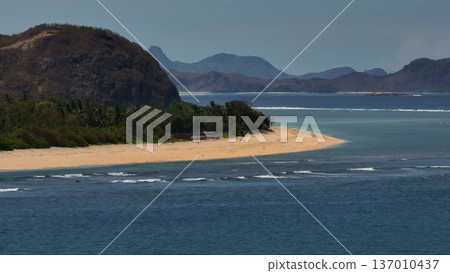 Lombok Pantai Tomang Omang beach coastline featuring a sandy shore meeting clear turquoise ocean waters, surrounded by lush tropical vegetation and distant mountains under a blue sky. Drone shot 137010437