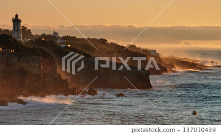 Portugal, Lisbon: Farol da Guia lighthouse rugged cliffs of Cascais coastline, golden sunset sunlight illuminate breaking waves and a small boat navigating the Atlantic Ocean. Dramatic landscape Portugal, Lisbon: Farol da Guia lighthouse rugged cliffs of Cascais coastline, golden sunset sunlight illuminate breaking waves and a small boat navigating the Atlantic Ocean. Dramatic landscape 137010439