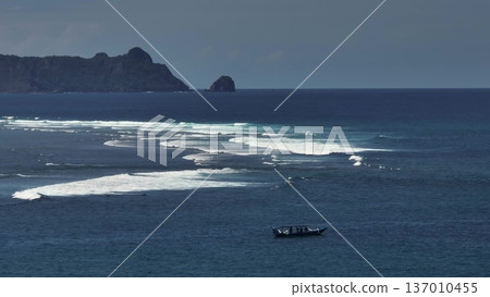 Lombok: Pantai Tomang Omang shoreline with a long white waves break of surf across deep blue sea, rugged island coastline in background and a small fishing boat on tranquil waters. Aerial drone flight Lombok: Pantai Tomang Omang shoreline with a long white waves break of surf across deep blue sea, rugged island coastline in background and a small fishing boat on tranquil waters. Aerial drone flight 137010455