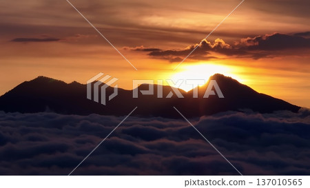 Mount Batur volcano silhouette at sunrise, with golden sunrays peaking over the mountain peak, creating a dramatic skyscape above a thick blanket of clouds. Travel to Bali, Indonesia. Aerial panorama 137010565