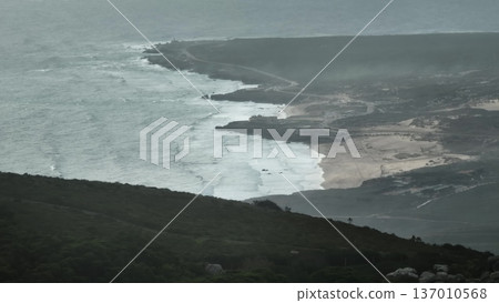 Portugal, Lisbon: Aerial capturing the rugged coastline of Sintra Cascais Natural Park, revealing powerful ocean waves crashing onto sandy beach and rocky shores under a misty, stormy sky 137010568
