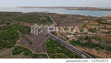 Malta: Cars and bus driving on a road crossing the countryside of Malta island, near the mediterranean sea, with Comino island visible in the background, aerial drone 137010573