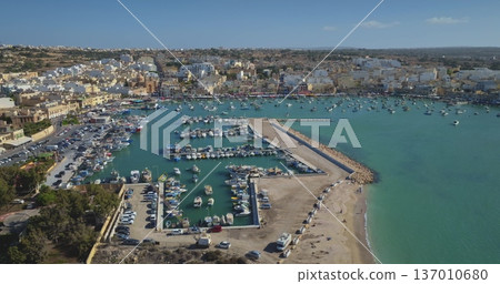 Malta: Aerial view capturing Marsaxlokk harbor in Malta coastline, showcasing colorful boats against backdrop of turquoise Mediterranean water, pier and buildings under blue sky. Drone flight Malta: Aerial view capturing Marsaxlokk harbor in Malta coastline, showcasing colorful boats against backdrop of turquoise Mediterranean water, pier and buildings under blue sky. Drone flight 137010680