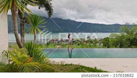 Indonesia, Bali: Woman enjoying tropical vacation, walking along edge of an infinity pool with stunning views of Lake Batur and surrounding green mountains in cloudy day. Relax, summer travel concept Indonesia, Bali: Woman enjoying tropical vacation, walking along edge of an infinity pool with stunning views of Lake Batur and surrounding green mountains in cloudy day. Relax, summer travel concept 137010682