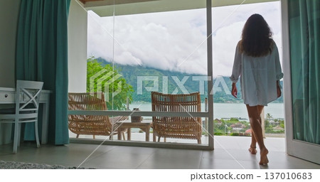 Indonesia, Bali, Batur: Woman in white shirt walk barefoot in modern hotel room to outdoor terrace with rattan chairs and table, enjoy beautiful mountain and lake landscape. Travel, holiday, vacation 137010683