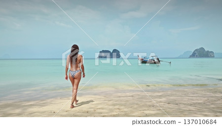 Thailand, Koh Lanta: Woman walking barefoot on sand tropical beach to turquoise water and traditional Thai longtail boat, distant limestone islands framing a tranquil vacation travel escape. Back view Thailand, Koh Lanta: Woman walking barefoot on sand tropical beach to turquoise water and traditional Thai longtail boat, distant limestone islands framing a tranquil vacation travel escape. Back view 137010684