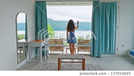 Beautiful woman in short dress standing in hotel room, looking in full-length window balcony at serene Batur lake and mountains, summer travel and relaxation in Indonesia, Bali. Slow motion, back view Beautiful woman in short dress standing in hotel room, looking in full-length window balcony at serene Batur lake and mountains, summer travel and relaxation in Indonesia, Bali. Slow motion, back view 137010685