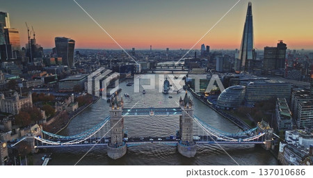 Aerial view Tower Bridge glowing over River Thames at sunset, evening traffic and boats framed by historic landmarks and modern skyscrapers. The Shard and Gherkin night cityscape. Drone flight footage 137010686