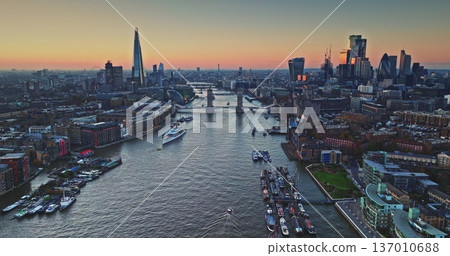 Evening aerial view of London cityscape during golden hour sunset, highlighting Tower Bridge spanning River Thames with The Shard and other iconic skyscrapers dominating the financial district skyline 137010688