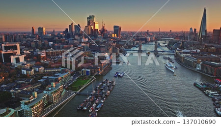 London sunset urban cityscape, financial district skyscrapers, Tower Bridge, The Shard reflecting warm evening light, boats navigating River Thames. Modern city skyline. Aerial drone flight panorama London sunset urban cityscape, financial district skyscrapers, Tower Bridge, The Shard reflecting warm evening light, boats navigating River Thames. Modern city skyline. Aerial drone flight panorama 137010690
