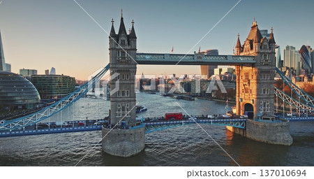 Tower Bridge Victorian Gothic towers and bustling cars traffic cross the River Thames at golden hour sunset, London skyline glowing in warm evening light. Travel destination. Aerial panorama footage Tower Bridge Victorian Gothic towers and bustling cars traffic cross the River Thames at golden hour sunset, London skyline glowing in warm evening light. Travel destination. Aerial panorama footage 137010694